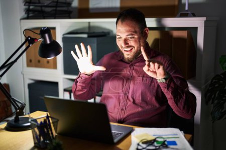 Foto de Hombre hispano de talla grande con barba trabajando en la oficina por la noche mostrando y señalando con los dedos el número seis mientras sonríe confiado y feliz. - Imagen libre de derechos