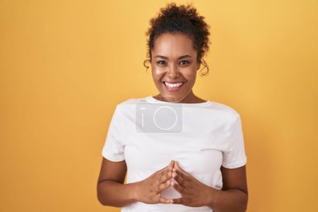 Photo for Young hispanic woman with curly hair standing over yellow background hands together and fingers crossed smiling relaxed and cheerful. success and optimistic - Royalty Free Image