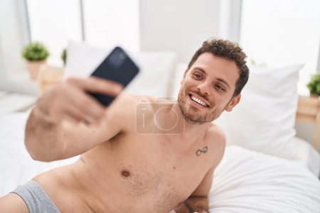 Foto de Hombre hispano joven haciendo selfie por el teléfono inteligente acostado en la cama en el dormitorio - Imagen libre de derechos
