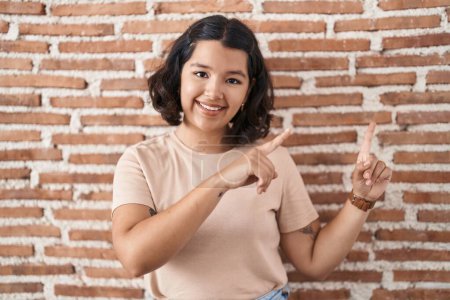 Foto de Mujer hispana joven de pie sobre la pared de ladrillos sonriendo y mirando a la cámara apuntando con dos manos y dedos hacia un lado. - Imagen libre de derechos