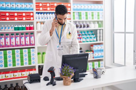 Téléchargez les photos : Jeune pharmacien arabe parlant sur smartphone en utilisant un ordinateur à la pharmacie - en image libre de droit