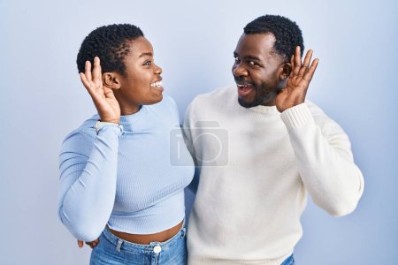 Foto de Joven pareja afroamericana de pie sobre fondo azul sonriendo con la mano sobre el oído escuchando un rumor o chismes. concepto de sordera. - Imagen libre de derechos