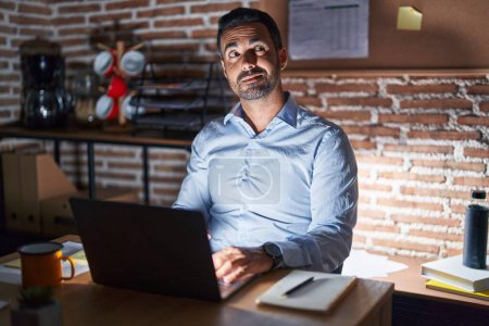Foto de Hombre hispano con barba trabajando en la oficina por la noche sonriendo mirando a un lado y mirando a otro pensando. - Imagen libre de derechos