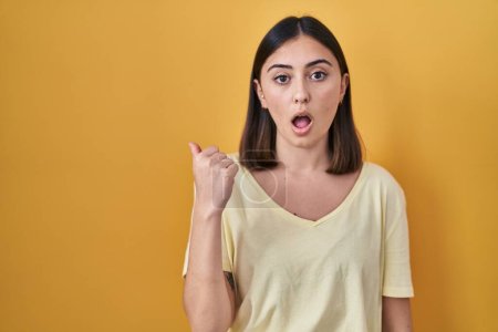 Photo for Hispanic girl wearing casual t shirt over yellow background surprised pointing with hand finger to the side, open mouth amazed expression. - Royalty Free Image