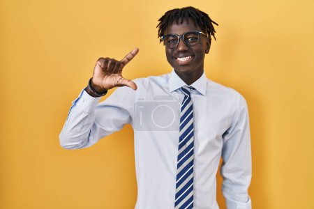 Foto de Hombre africano con rastas de pie sobre fondo amarillo sonriente y seguro gesto con la mano haciendo signo de pequeño tamaño con los dedos mirando y la cámara. concepto de medida. - Imagen libre de derechos