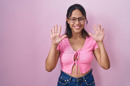 Téléchargez les photos : Jeune femme hispanique debout sur fond rose portant des lunettes montrant et pointant vers le haut avec les doigts numéro dix tout en souriant confiant et heureux. - en image libre de droit