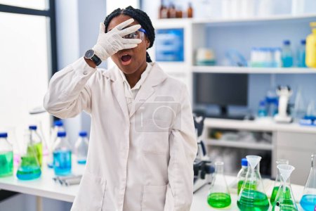 Foto de Mujer afroamericana trabajando en laboratorio científico asomándose en shock cubriendo cara y ojos con la mano, mirando a través de los dedos con miedo - Imagen libre de derechos