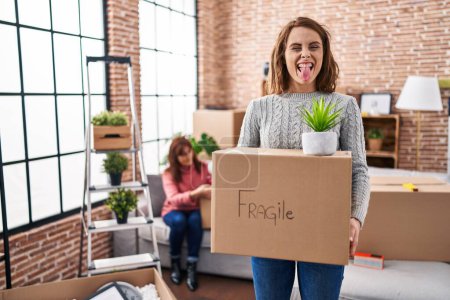 Photo for Mother and daughter moving to a new home holding cardboard box sticking tongue out happy with funny expression. - Royalty Free Image
