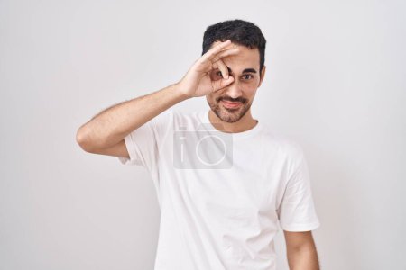 Téléchargez les photos : Bel homme hispanique debout sur fond blanc faisant un geste correct avec la main souriante, les yeux regardant à travers les doigts avec un visage heureux. - en image libre de droit