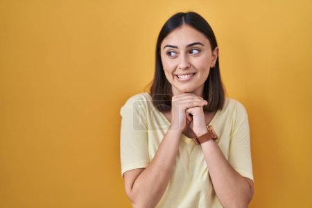 Foto de Chica hispana vistiendo camiseta casual sobre fondo amarillo riendo nerviosa y emocionada con las manos en la barbilla mirando a un lado - Imagen libre de derechos