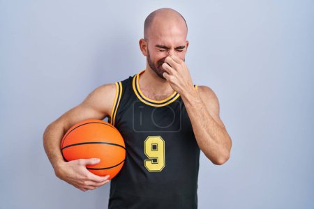 Téléchargez les photos : Jeune homme chauve avec barbe portant un uniforme de basket tenant la balle sentant quelque chose de puant et dégoûtant, odeur intolérable, retenant la respiration avec les doigts sur le nez. mauvaise odeur - en image libre de droit