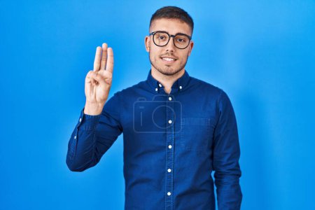 Photo for Young hispanic man wearing glasses over blue background showing and pointing up with fingers number three while smiling confident and happy. - Royalty Free Image