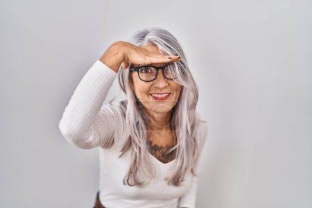 Foto de Mujer de mediana edad con el pelo gris de pie sobre fondo blanco muy feliz y sonriente mirando lejos con la mano sobre la cabeza. concepto de búsqueda. - Imagen libre de derechos