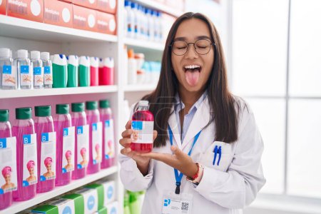 Téléchargez les photos : Young hispanic woman working at pharmacy drugstore holding syrup sticking tongue out happy with funny expression. - en image libre de droit