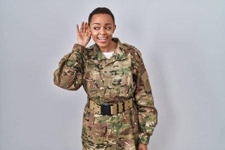 Foto de Hermosa mujer afroamericana vistiendo uniforme camuflaje del ejército sonriendo con la mano sobre la oreja escuchando un rumor o chismes. concepto de sordera. - Imagen libre de derechos