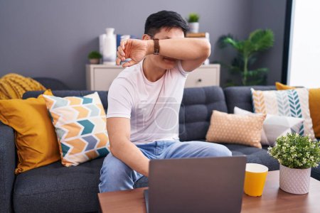 Téléchargez les photos : Jeune homme asiatique à l'aide d'un ordinateur portable à la maison assis sur le canapé souriant gai jouer jeter un coup d'oeil un boo avec les mains montrant le visage. surpris et sortis - en image libre de droit