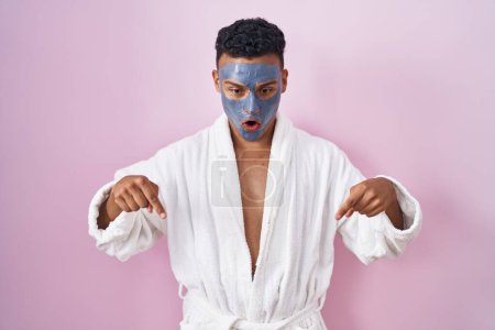 Téléchargez les photos : Jeune homme hispanique portant un masque de beauté et peignoir de bain pointant vers le bas avec les doigts montrant la publicité, visage surpris et bouche ouverte - en image libre de droit