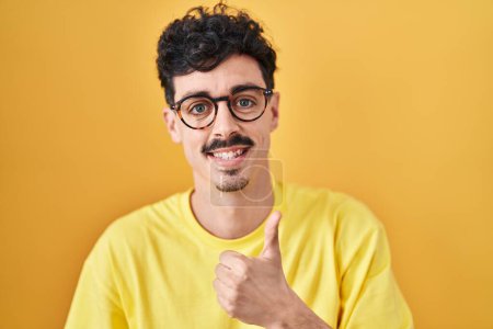 Foto de Hombre hispano con gafas de pie sobre fondo amarillo haciendo un gesto feliz con la mano. aprobando la expresión mirando a la cámara mostrando éxito. - Imagen libre de derechos
