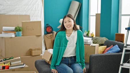 Téléchargez les photos : Jeune femme chinoise souriant confiant assis sur le canapé à la nouvelle maison - en image libre de droit