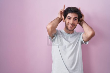 Téléchargez les photos : Jeune homme hispanique debout sur fond rose posant drôle et fou avec les doigts sur la tête comme des oreilles de lapin, souriant joyeux - en image libre de droit
