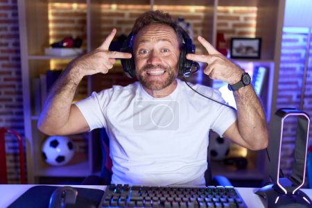 Photo for Middle age man with beard playing video games wearing headphones doing peace symbol with fingers over face, smiling cheerful showing victory - Royalty Free Image