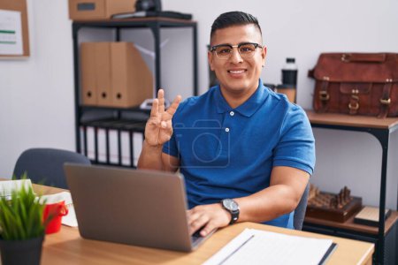 Foto de Joven hombre hispano trabajando en la oficina con portátil mostrando y señalando con los dedos número tres mientras sonríe confiado y feliz. - Imagen libre de derechos