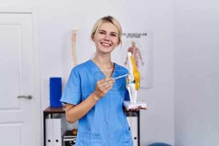 Foto de Joven mujer rubia pisioterapeuta sonriendo confiada apuntando a modelo anatómico de rodilla en clínica de rehabilitación - Imagen libre de derechos