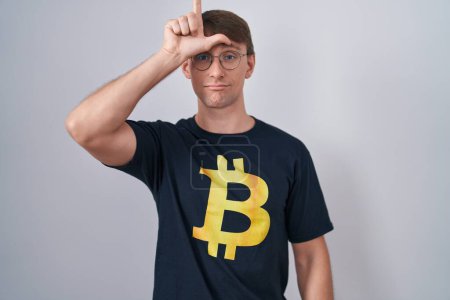 Photo for Caucasian blond man wearing bitcoin t shirt making fun of people with fingers on forehead doing loser gesture mocking and insulting. - Royalty Free Image