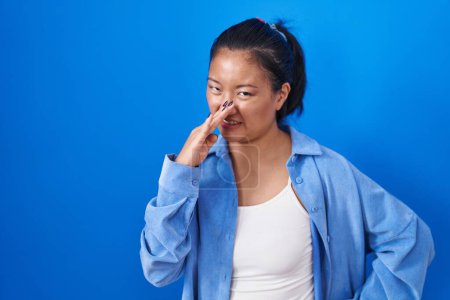 Téléchargez les photos : Jeune femme asiatique debout sur fond bleu sentant quelque chose de puant et dégoûtant, odeur intolérable, retenant souffle avec les doigts sur le nez. mauvaise odeur - en image libre de droit