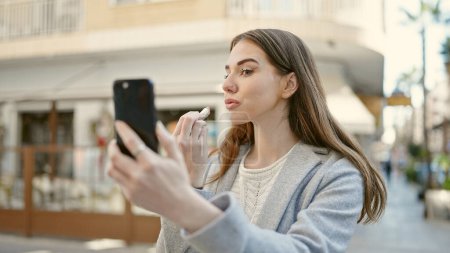 Téléchargez les photos : Jeune femme hispanique utilisant le smartphone comme miroir appliquant du rouge à lèvres sur la terrasse du café - en image libre de droit