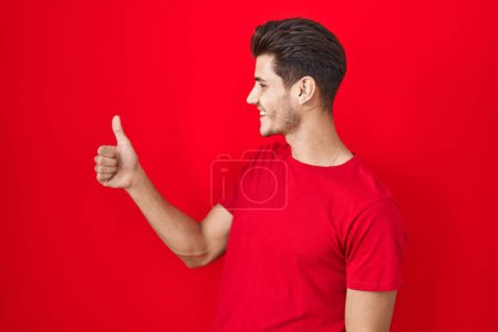 Photo for Young hispanic man standing over red background looking proud, smiling doing thumbs up gesture to the side - Royalty Free Image