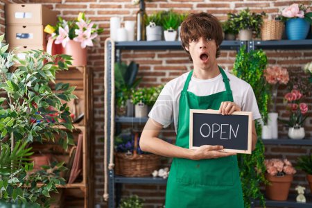 Photo for Hispanic young man working at florist holding open sign in shock face, looking skeptical and sarcastic, surprised with open mouth - Royalty Free Image