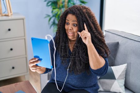Photo for Plus size hispanic woman using touchpad wearing headphones smiling with an idea or question pointing finger with happy face, number one - Royalty Free Image