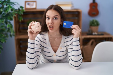 Foto de Joven chica hispana sosteniendo alcancía y tarjeta de crédito expresión despistada y confusa. concepto de duda. - Imagen libre de derechos