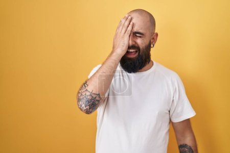 Photo for Young hispanic man with beard and tattoos standing over yellow background yawning tired covering half face, eye and mouth with hand. face hurts in pain. - Royalty Free Image