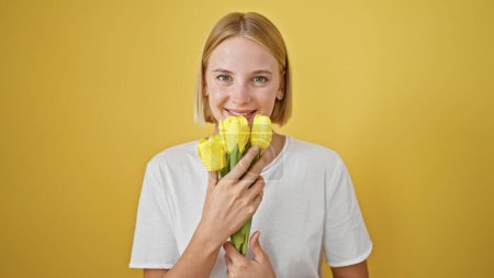 Photo for Young blonde woman smelling flowers smiling over isolated yellow background - Royalty Free Image