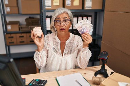 Photo for Middle age woman with grey hair working at small business ecommerce holding piggy bank and zloty clueless and confused expression. doubt concept. - Royalty Free Image