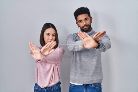 Foto de Joven pareja hispana de pie junta expresión de rechazo cruzando brazos y palmas haciendo signo negativo, cara enojada - Imagen libre de derechos