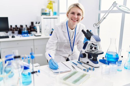 Photo for Young blonde woman scientist using microscope taking notes at laboratory - Royalty Free Image