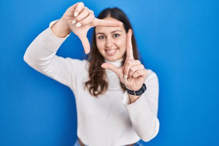 Téléchargez les photos : Jeune femme hispanique debout sur fond bleu souriant faisant cadre avec les mains et les doigts avec le visage heureux. concept de créativité et photographie. - en image libre de droit