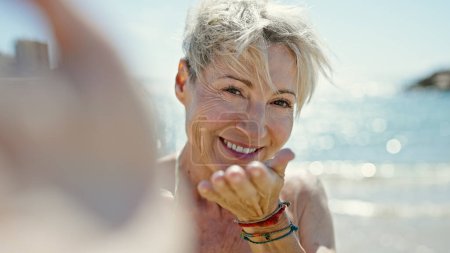 Téléchargez les photos : Moyen âge femme blonde touriste portant un bikini faire selfie souffler baiser à la plage - en image libre de droit
