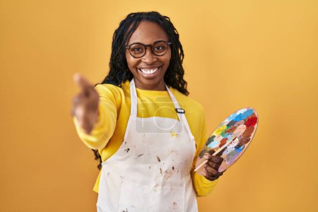 Téléchargez les photos : Femme africaine tenant palette peintre souriant amical offrant poignée de main comme salutation et accueil. entreprise prospère. - en image libre de droit