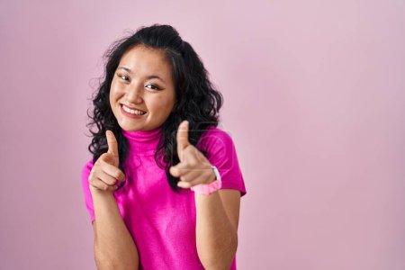 Foto de Joven mujer asiática de pie sobre fondo rosa señalando los dedos a la cámara con la cara feliz y divertida. buena energía y vibraciones. - Imagen libre de derechos