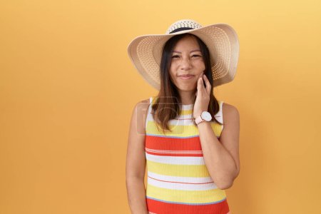 Téléchargez les photos : Femme chinoise d'âge moyen portant un chapeau d'été sur fond jaune touchant la bouche avec la main avec une expression douloureuse en raison de maux de dents ou de maladies dentaires sur les dents. dentiste - en image libre de droit