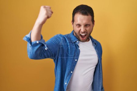 Photo for Hispanic man standing over yellow background angry and mad raising fist frustrated and furious while shouting with anger. rage and aggressive concept. - Royalty Free Image