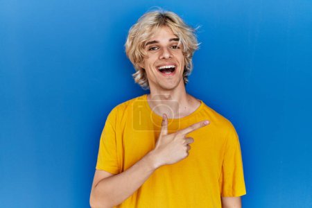 Téléchargez les photos : Jeune homme moderne debout sur fond bleu joyeux avec un sourire du visage pointant avec la main et le doigt sur le côté avec une expression heureuse et naturelle sur le visage - en image libre de droit