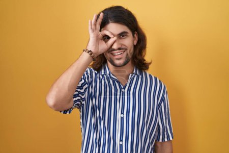 Photo for Hispanic man with long hair standing over yellow background doing ok gesture with hand smiling, eye looking through fingers with happy face. - Royalty Free Image