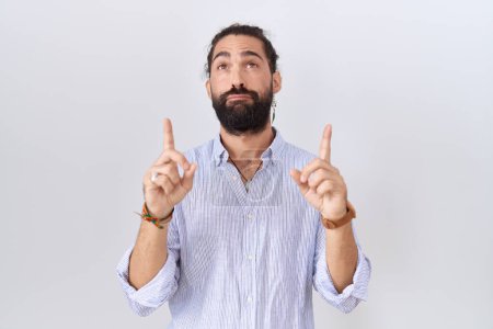 Foto de Hombre hispano con barba llevando camisa casual apuntando hacia arriba mirando triste y molesto, indicando dirección con los dedos, infeliz y deprimido. - Imagen libre de derechos