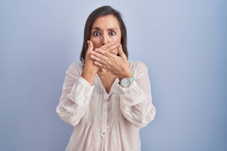 Foto de Mujer hispana de mediana edad de pie sobre fondo azul sorprendió cubriendo la boca con las manos por error. concepto secreto. - Imagen libre de derechos