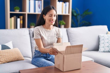 Photo for Young hispanic woman opening cardboard box winking looking at the camera with sexy expression, cheerful and happy face. - Royalty Free Image